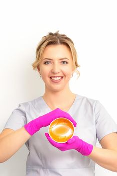 Portrait of a female caucasian beautician holding a jar of sugar paste for sugaring wearing pink gloves on white background