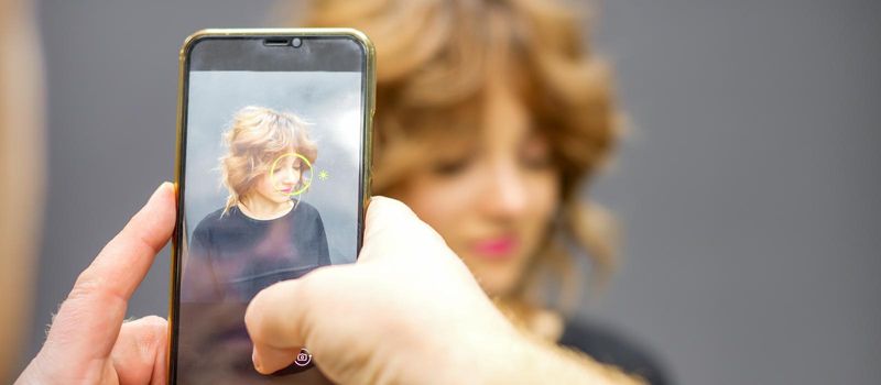 Man hairdresser taking pictures on the smartphone of her client's hairstyle against a gray background