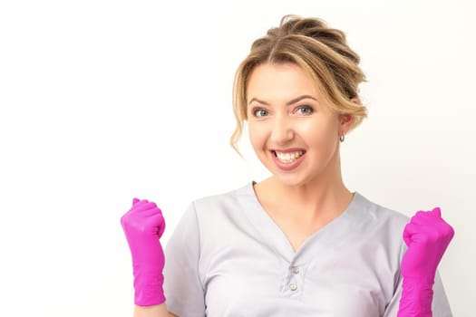 Happy caucasian woman doctor wearing pink gloves celebrates and raising fists on white background