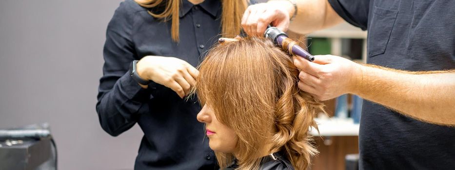 Two hairstylists make curls hairstyle of long brown hair with the curling iron in hairdresser salon