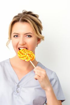 Portrait of a beautiful young caucasian beautician wearing a medical shirt bites a lollipop on a white background