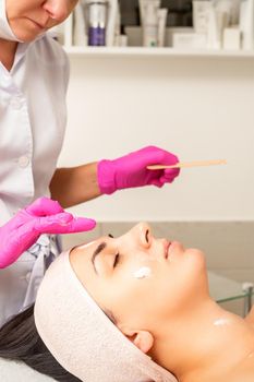 Cosmetologist with gloved hands applies a moisturizing mask with peeling cream on the female face. Facial cosmetology treatment. Procedures for facial care