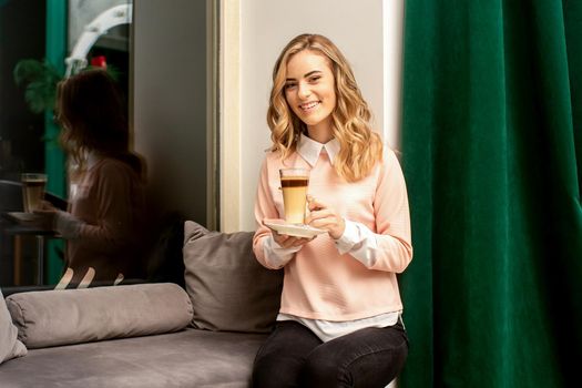 Beautiful caucasian young smiling woman with long wavy blonde hair holding and drinking a latte from a glass cup, sitting on a sofa