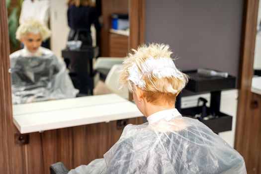 Coloring white hair with hair dye of the young caucasian blonde woman sitting at a hair salon, close up