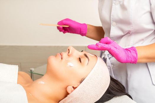 Cosmetologist with gloved hands applies a moisturizing mask with peeling cream on the female face. Facial cosmetology treatment. Procedures for facial care