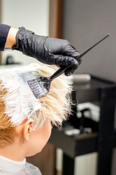 Coloring white hair with hair dye and brush by hands of hairstylist for the young caucasian blonde woman at a hair salon, close up