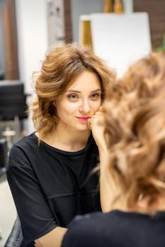 The beautiful young caucasian red-haired woman with a new short wavy hairstyle looking in the mirror checking hairstyle and makeup in a hairdresser salon