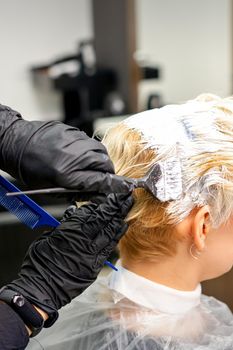 Coloring white hair with hair dye and brush by hands of hairstylist for the young caucasian blonde woman at a hair salon, close up