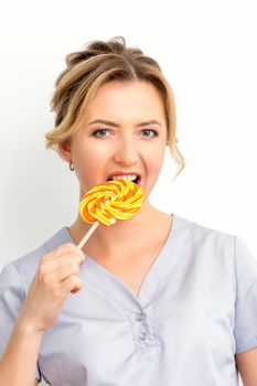 Portrait of a beautiful young caucasian beautician wearing a medical shirt bites a lollipop on a white background