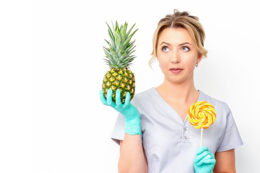 Young caucasian female doctor nutritionist holding fresh pineapple with lollipop over white background