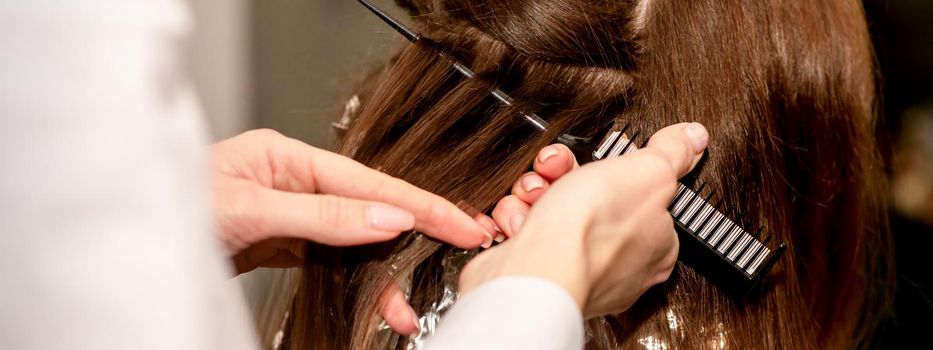 Hairdresser's hands prepare brown hair for dyeing with a comb and foil in a beauty salon, close up