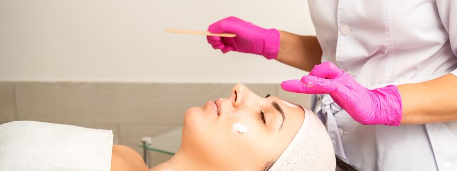 Cosmetologist with gloved hands applies a moisturizing mask with peeling cream on the female face. Facial cosmetology treatment. Procedures for facial care