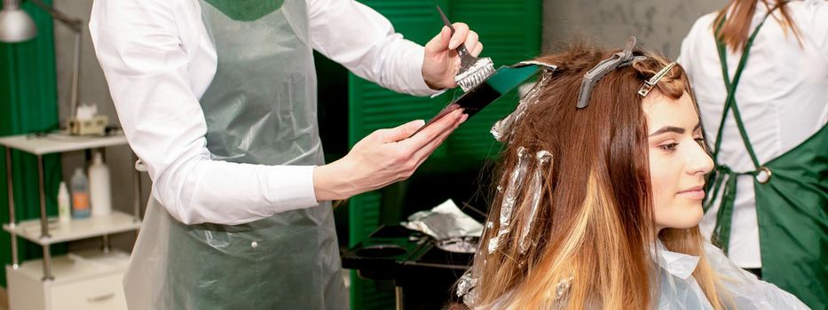 A female hairdresser dyes the hair of a young Caucasian woman with a brush and foil in a beauty salon