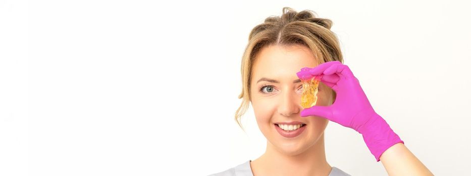 The master of sugar hair removal holds liquid yellow sugar paste, covering her eye with wax for depilation on a white background