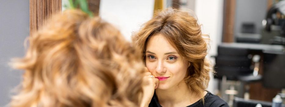 The beautiful young caucasian red-haired woman with a new short wavy hairstyle looking in the mirror checking hairstyle and makeup in a hairdresser salon