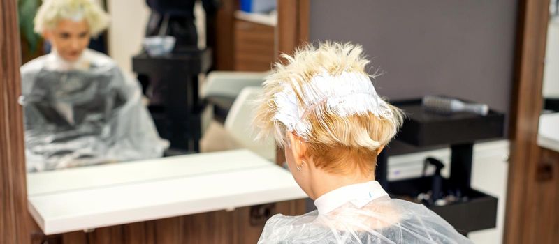 Coloring white hair with hair dye of the young caucasian blonde woman sitting at a hair salon, close up