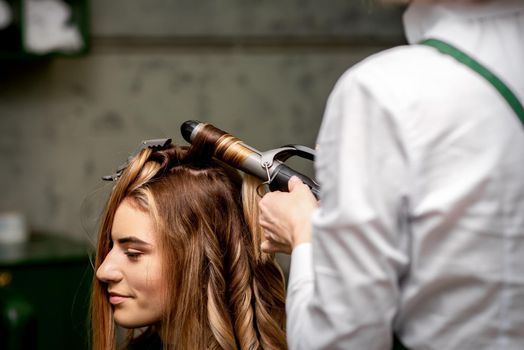 The female hairdresser is curling hair for a brown-haired young caucasian woman in a beauty salon