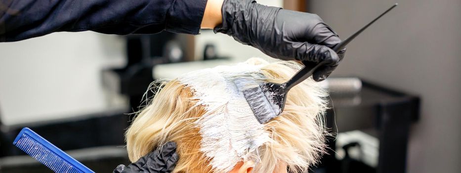 Coloring white hair with hair dye and brush by hands of hairstylist for the young caucasian blonde woman at a hair salon, close up