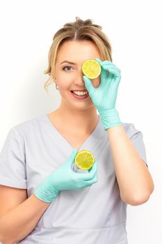 Portrait of young caucasian smiling female beautician covering eye with a lime slice wearing gloves over a white background