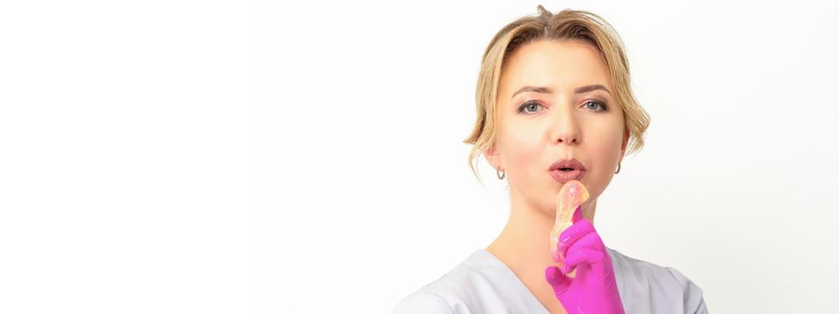 Young caucasian female beautician holding wax paste on forefinger blowing on him wearing pink gloves against a white background. Epilation concept