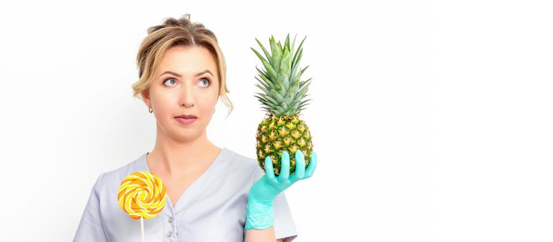 Young caucasian female doctor nutritionist holding fresh pineapple with lollipop over white background