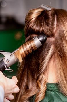 The female hairdresser is curling hair for a brown-haired young caucasian woman in a beauty salon