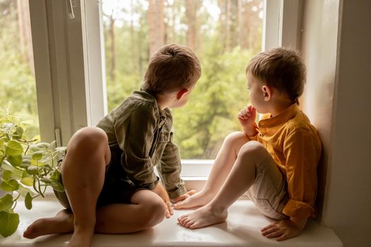 Children sitting on windowsill and waiting for someone comming. Two brothers, friends. Cute preschool kids alone at home. Boys are waiting for their mother or father. Loneliness. Busy parents