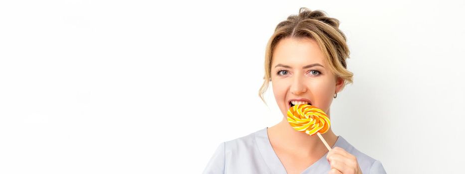 Portrait of a beautiful young caucasian beautician wearing a medical shirt bites a lollipop on a white background