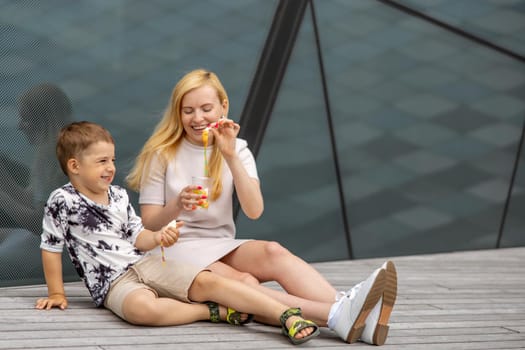 Happy blond woman and little boy sitting on terrace and eating sweets. Mother and son enjoy time together. Positive young mom playing, spending time with her cute child, laughing, having fun. Family