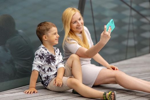 Happy blond woman and little boy sitting on terrace and making selfie on smartphone. Mother and son enjoy time together. Positive young mom spending time with her cute child. Having fun. Family