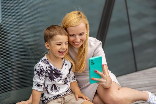 Happy blond woman and little boy sitting on terrace and making selfie on smartphone. Mother and son enjoy time together. Positive young mom spending time with her cute child. Having fun. Family
