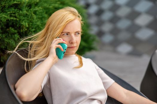 Young blond woman sittings outdoors in the city and making a call with her smartphone. Girl talking with friends with mobile phone. Leisure activity, communication. Conversation. Mobile technology