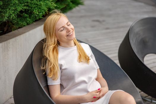 Young beautiful blond woman sitting outdoors, on the wooden terrace in the city and resting. Girl has break, spending time outside, relaxing. Time with yourself, dreaming, relaxation, mental health