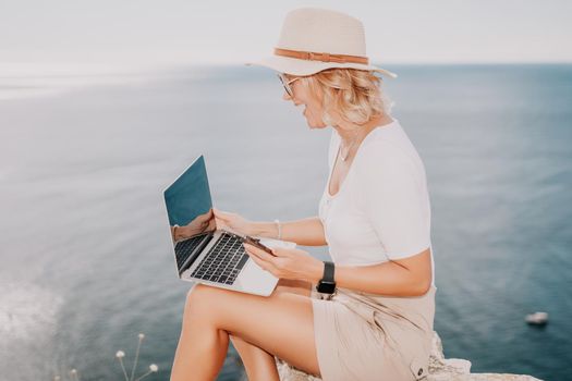 Successful business woman in yellow hat working on laptop by the sea. Pretty lady typing on computer at summer day outdoors. Freelance, travel and holidays concept.
