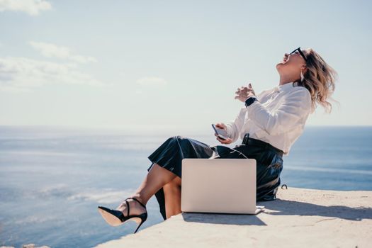 Successful business woman in yellow hat working on laptop by the sea. Pretty lady typing on computer at summer day outdoors. Freelance, travel and holidays concept.