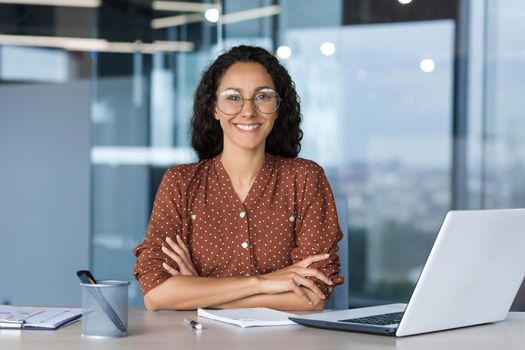 Portrait of successful young businesswoman, Hispanic woman working inside modern office building smiling and looking at camera sitting at desk with laptop, female worker with curly hair and glasses