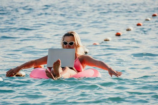Freelancer, young blond woman in sunglases floating on an inflatable big pink donut with a laptop in the sea at sunset. Freelance, travel and holidays concept. Slow motion