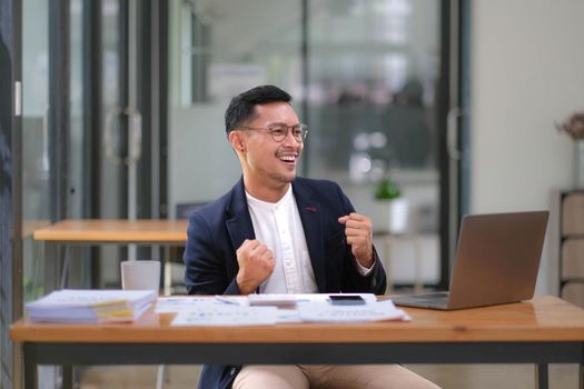 Portrait of an Asian male business owner standing with a computer showing happiness after a successful investment..