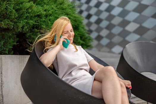 Young blond woman sittings outdoors in the city and making a call with her smartphone. Girl talking with friends with mobile phone. Leisure activity, communication. Conversation. Mobile technology