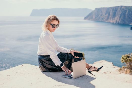 Successful business woman in yellow hat working on laptop by the sea. Pretty lady typing on computer at summer day outdoors. Freelance, travel and holidays concept.