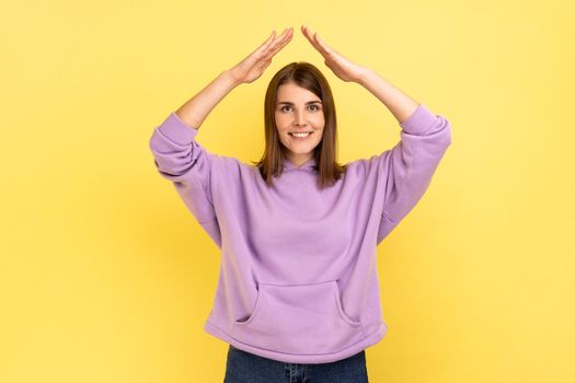 I'm in safety. Portrait of happy protected woman raising hands above head, making roof gesture, insurance, wearing purple hoodie. Indoor studio shot isolated on yellow background.