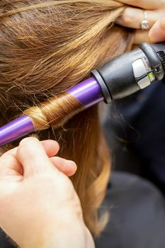 The hairstylist makes curls hairstyle of long brown hair with the curling iron in hairdresser salon, close up