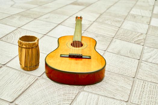 a stringed musical instrument, with a fretted fingerboard. Yellow acoustic guitar and straw decorative keg on gray checkered