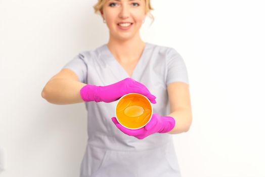 Portrait of a female caucasian beautician holding a jar of sugar paste for sugaring wearing pink gloves on white background