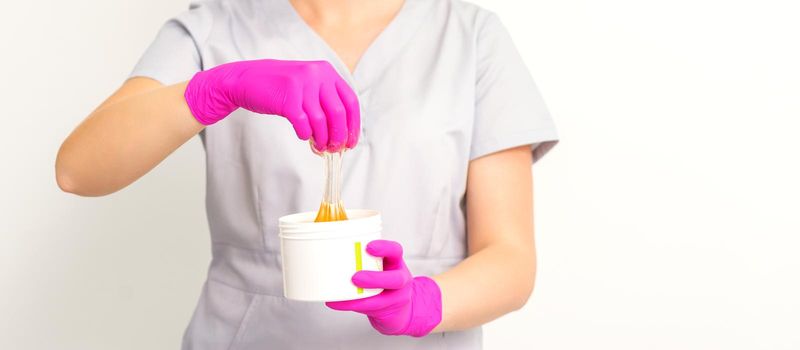 Portrait of a female caucasian beautician holding a jar of sugar paste for sugaring wearing pink gloves on white background