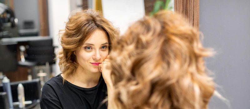 The beautiful young caucasian red-haired woman with a new short wavy hairstyle looking in the mirror checking hairstyle and makeup in a hairdresser salon