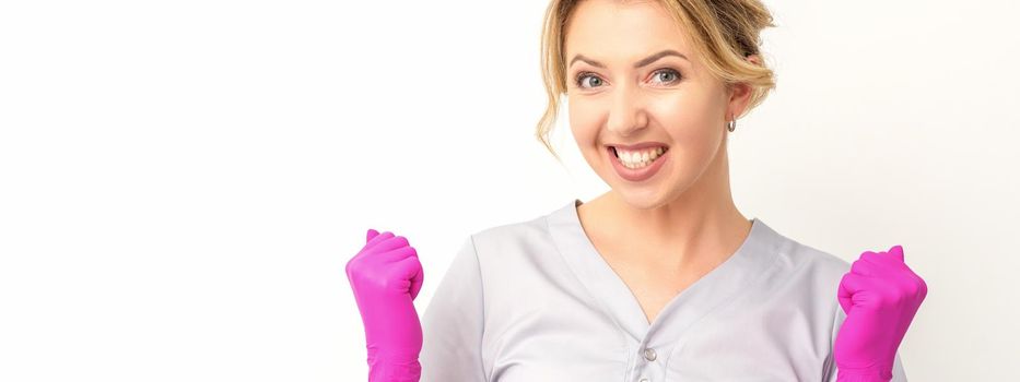 Happy caucasian woman doctor wearing pink gloves celebrates and raising fists on white background