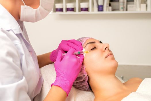 The young white woman is getting rejuvenating facial injections with hyaluronic acid on the eye in a beauty clinic