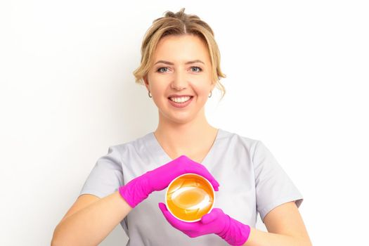 Portrait of a female caucasian beautician holding a jar of sugar paste for sugaring wearing pink gloves on white background