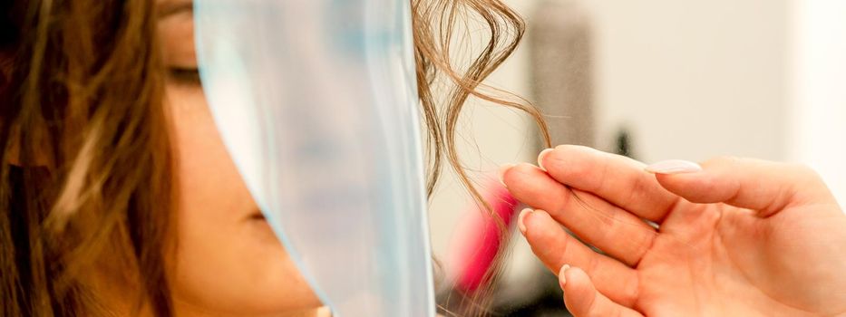 Professional hairdresser styling and fixing with hairspray long woman curly hair covering the face with a protective shield in a beauty salon